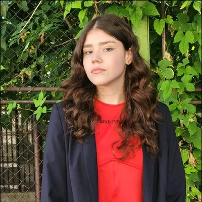 Ana Farazică, in een rood shirt en donker jasje, voor groene bladeren.