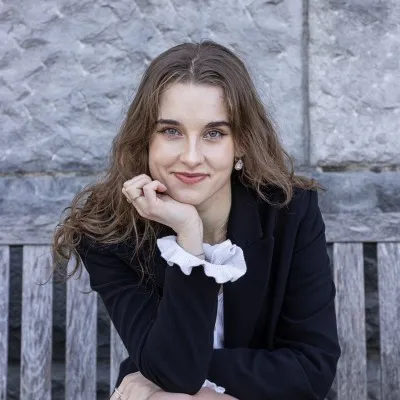 Kinga Marchlewska, met haar kin op haar hand, in een donkere blazer over een witte blouse met ruches, op een houten bankje voor een stenen muur.